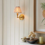 Endon Ashstead LED wall light with brushed brass arm and pleated white fabric shade, lit on a panelled wall beside a framed pineapple print.