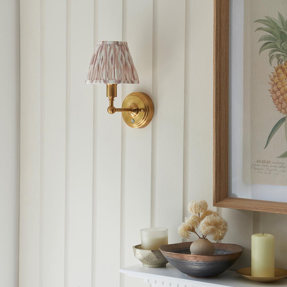Endon Ashstead LED Ikat brushed brass wall light with pleated pink and natural fabric shade on a cream panelled wall beside a framed pineapple print.