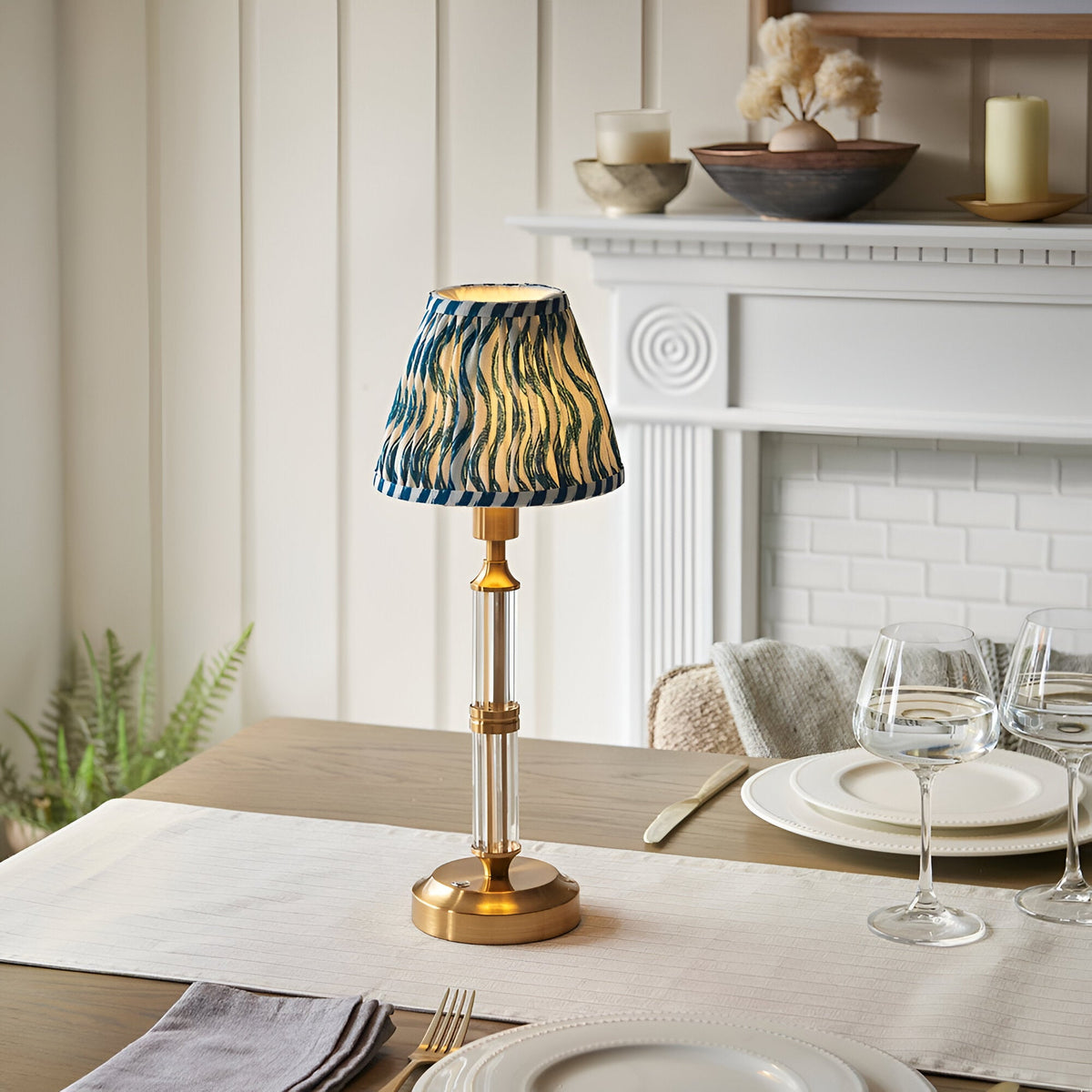 Endon Carlwood LED table lamp with brushed brass base, clear acrylic detail and blue Ripple pleated shade, on a dining table by a white fireplace.