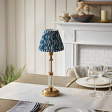 Endon Carlwood rechargeable LED table lamp in brushed brass with clear acrylic stem and blue Ripple pleated shade, on a dining table by a white mantel.