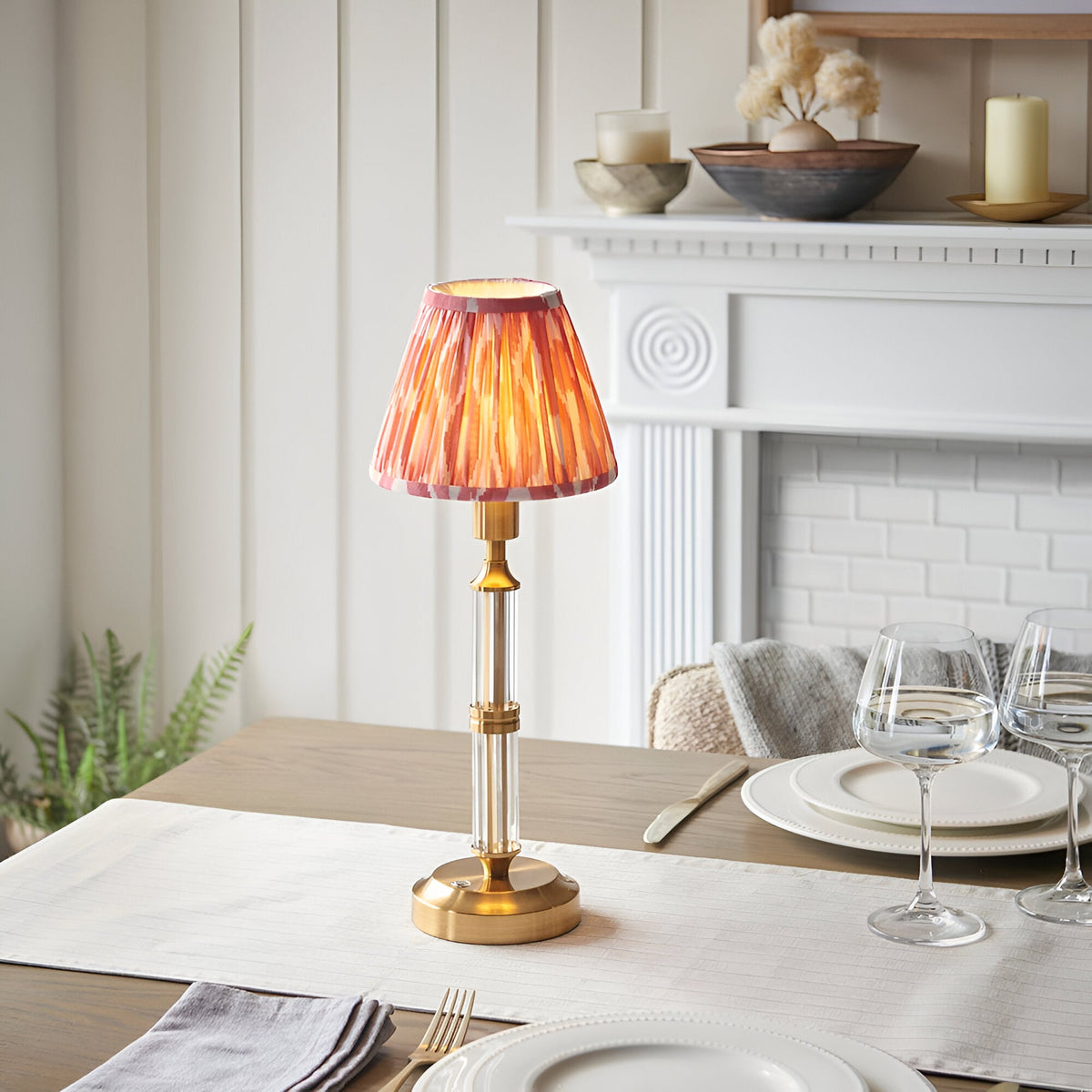 Endon Carlwood LED table lamp with brushed brass base, clear acrylic stem and pleated coral pink Ikat shade, lit on a dining table by a white fireplace.