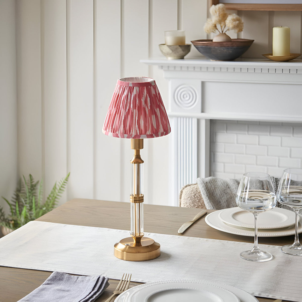 Endon Carlwood cordless LED table lamp with brushed brass base, clear acrylic detail and pink Ikat pleated shade on a dining table by a white fireplace.