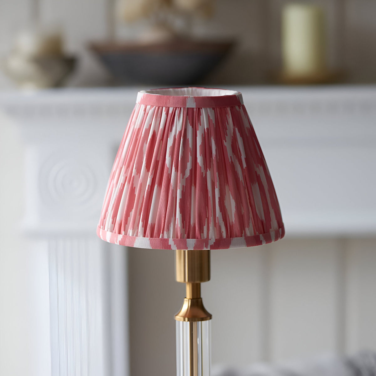 Endon Carlwood LED table lamp with a pleated coral pink and white Ikat shade on a brushed brass base, shown on a mantel setting.