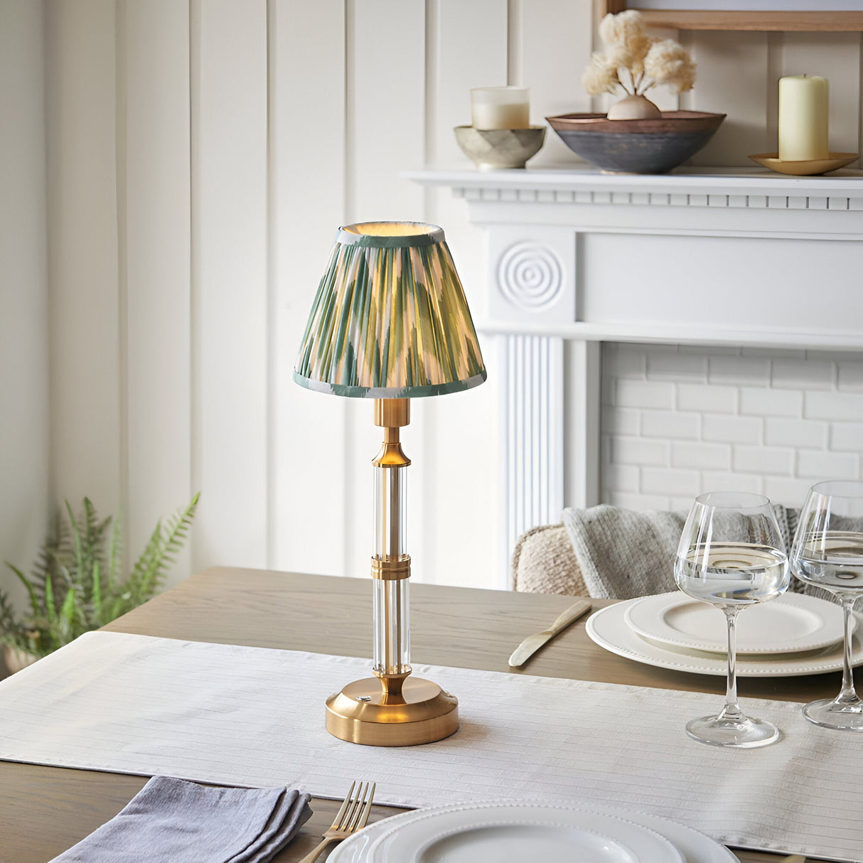 Carlwood LED Table Lamp in brushed aged brass with clear acrylic column and pleated green Zigzag shade, shown on a dining table near a white mantelpiece.