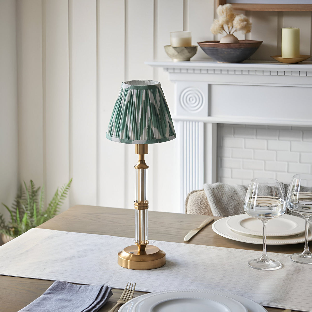 Carlwood LED table lamp with brushed aged brass base, clear acrylic column, and pleated Cotswold green zigzag shade on a dining table.
