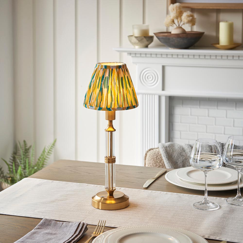 Carlwood LED table lamp with brass column, glass stem, and green-yellow Ikat fabric shade, shown lit on a dining table near a white fireplace.