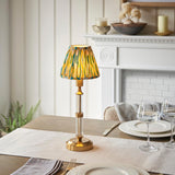 Carlwood LED table lamp with brass column, glass stem, and green-yellow Ikat fabric shade, shown lit on a dining table near a white fireplace.