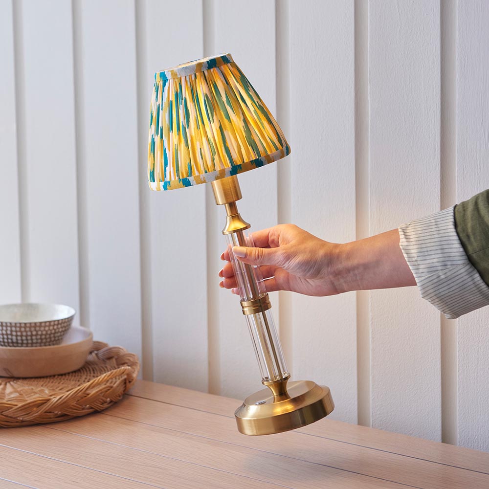 Carlwood LED table lamp with brass base, clear glass detail, and pleated yellow, teal and white Ikat shade on a wooden sideboard.