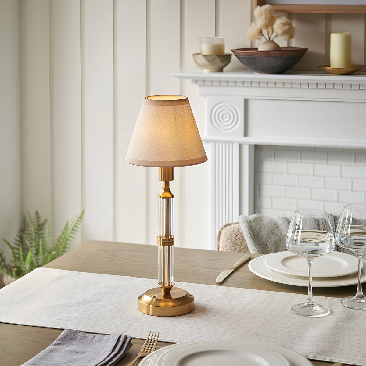 Endon Carlwood LED rechargeable table lamp in brushed brass with clear acrylic stem and vintage white tapered shade, set on a dining table by a fireplace.