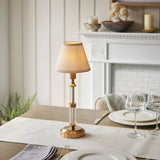 Endon Carlwood LED rechargeable table lamp in brushed brass with clear acrylic stem and vintage white tapered shade, set on a dining table by a fireplace.