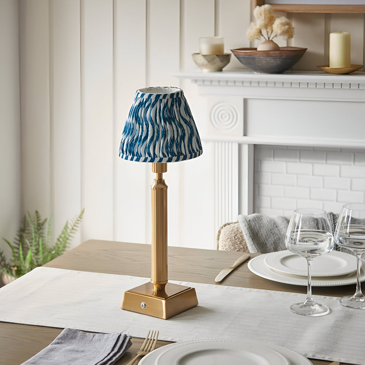 Cranwick LED Rechargeable Ripple Table Lamp with reeded brass stem, square base and blue-white patterned fabric shade, shown on a dining table near a fireplace.