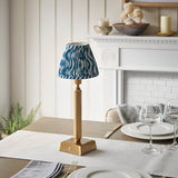 Cranwick LED Rechargeable Ripple Table Lamp with reeded brass stem, square base and blue-white patterned fabric shade, shown on a dining table near a fireplace.