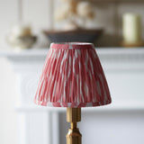 Endon Cranwick LED Ikat table lamp with a pleated pink and white shade on a brushed brass stem, set before a white fireplace mantel.