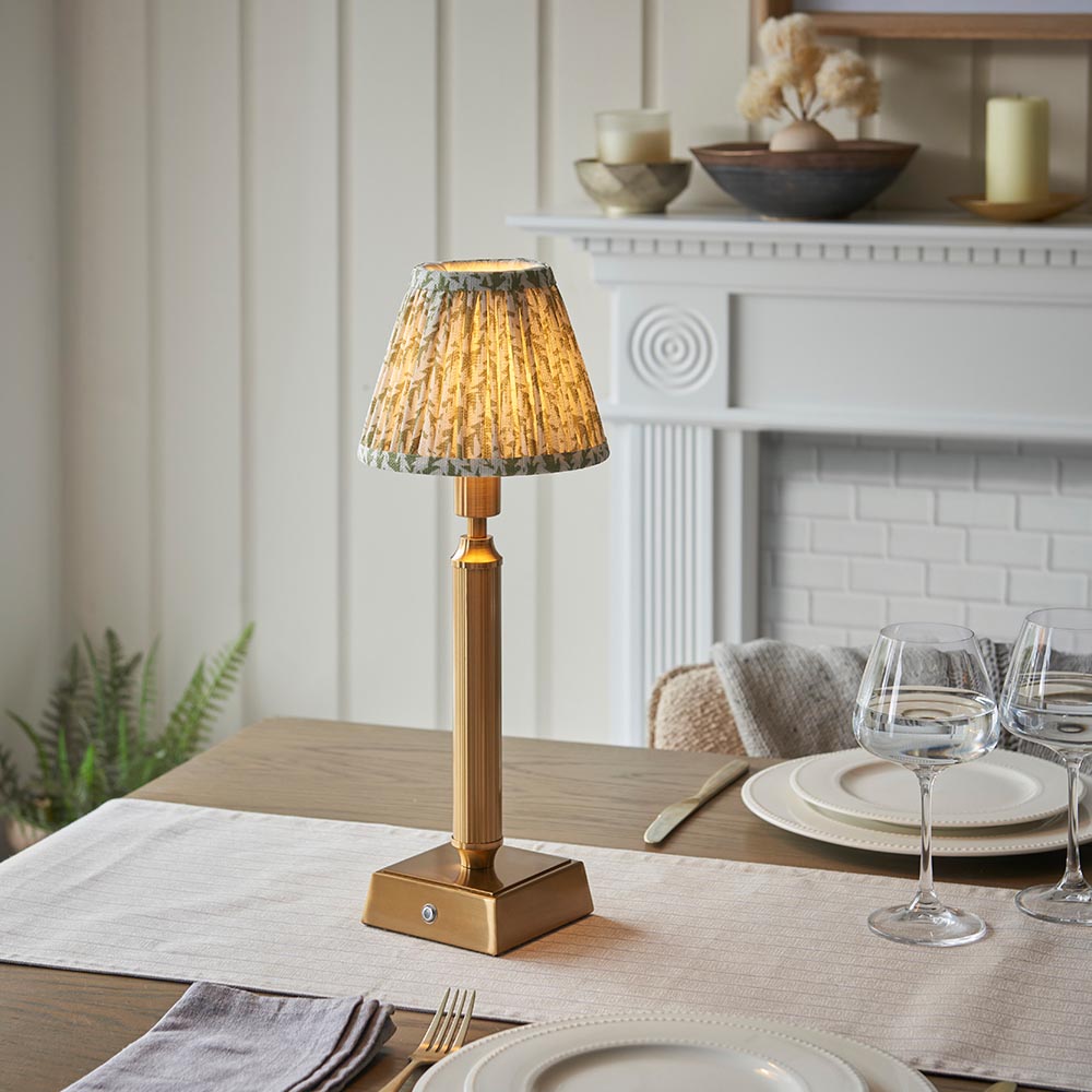 Endon Cranwick LED Leaf table lamp with brushed brass reeded stem and pleated green leaf-print shade on a dining table set with glassware.