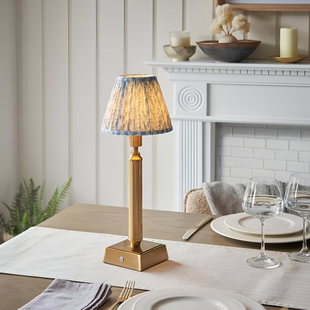 Endon Cranwick LED table lamp with brushed brass reeded stem and blue leaf-print pleated shade, on a dining table beside wine glasses and a white fireplace.