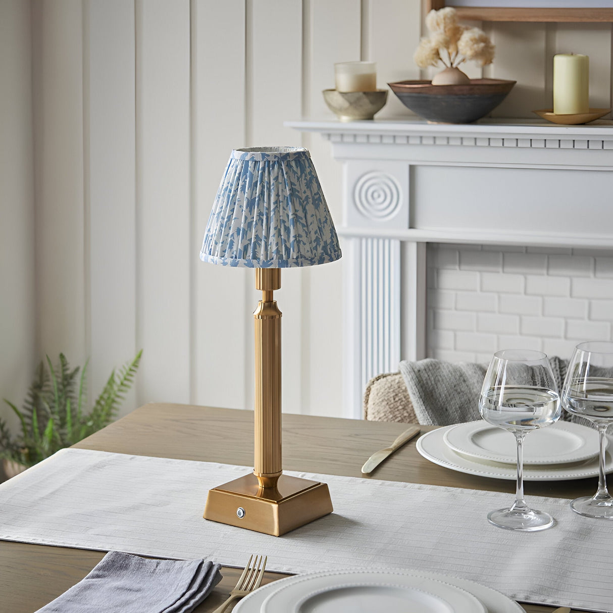 Endon Cranwick LED table lamp with reeded brushed brass stem and pleated blue leaf-print shade, on a dining table beside a white fireplace.