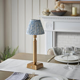 Endon Cranwick LED table lamp with reeded brushed brass stem and pleated blue leaf-print shade, on a dining table beside a white fireplace.
