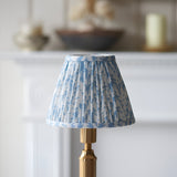 Endon Cranwick LED Leaf table lamp with blue and white pleated faux linen shade on a brushed brass reeded stem, shown on a mantelpiece.