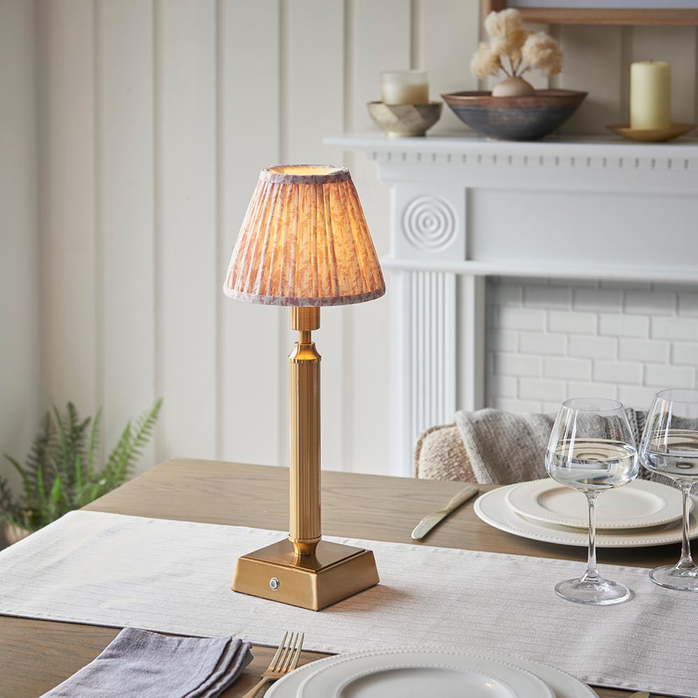 Cranwick LED Leaf Table Lamp in brass with a pleated fabric shade, shown lit on a dining table beside a white fireplace mantel.