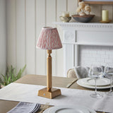 Cranwick LED Leaf Table Lamp in brass with a pink leaf-print fabric shade on a reeded column, shown on a dining table beside glassware and a fireplace.