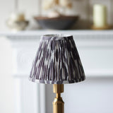 Endon Cranwick LED Ikat table lamp with brushed brass reeded stem and grey and white pleated Ikat shade, shown by a white fireplace mantel.