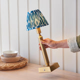 Cranwick LED Ikat Table Lamp with brass reeded stem, square base and blue patterned fabric shade, held by hand in a room setting.