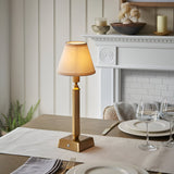 Endon Cranwick LED rechargeable table lamp with reeded brushed brass stem and ivy white fabric shade, lit on a dining table beside a white fireplace.