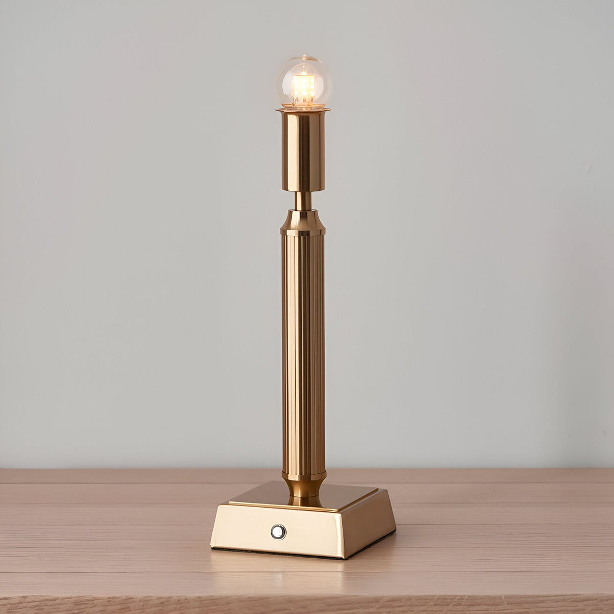 Endon Cranwick rechargeable LED table lamp lit on a wooden surface, showing its reeded brass stem, square base with touch button, and clear globe bulb.