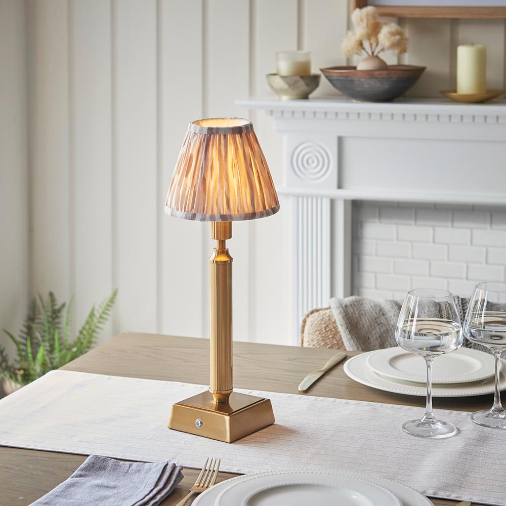 Cranwick LED Ikat table lamp with brass reeded stem, square base and pleated natural fabric shade, shown lit on a dining table near a fireplace.