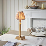 Cranwick LED Ikat table lamp with brass reeded stem, square base and pleated natural fabric shade, shown lit on a dining table near a fireplace.