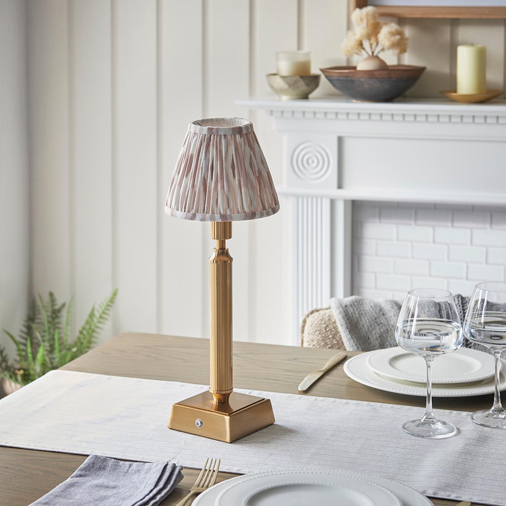 Cranwick LED Ikat Table Lamp with brass reeded stem, square base and patterned fabric shade, shown on a dining table near a white fireplace.