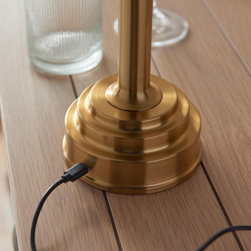 Close-up of the Endon Danebourne LED Leaf table lamp's stepped brushed brass base with USB-C charging cable connected, on a wooden slatted surface.