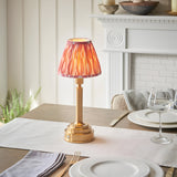 Endon Danebourne LED table lamp with brushed brass stepped base and pink Ikat pleated shade, on a dining table set with glassware and cutlery.