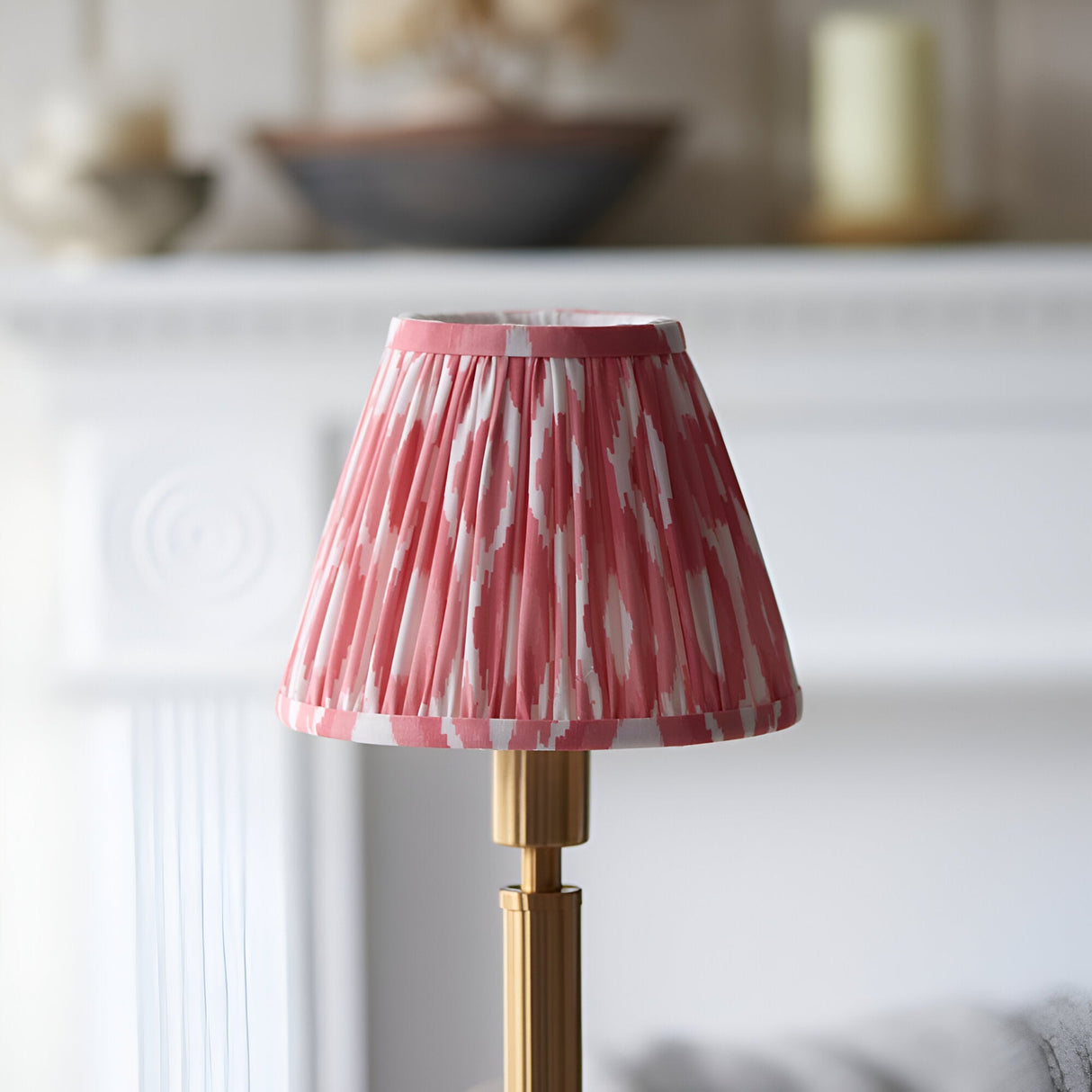 Endon Danebourne LED table lamp with a pleated pink and white ikat shade on a brushed brass stepped base, shown on a white mantelpiece.