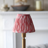 Endon Danebourne LED table lamp with a pleated pink and white ikat shade on a brushed brass stepped base, shown on a white mantelpiece.