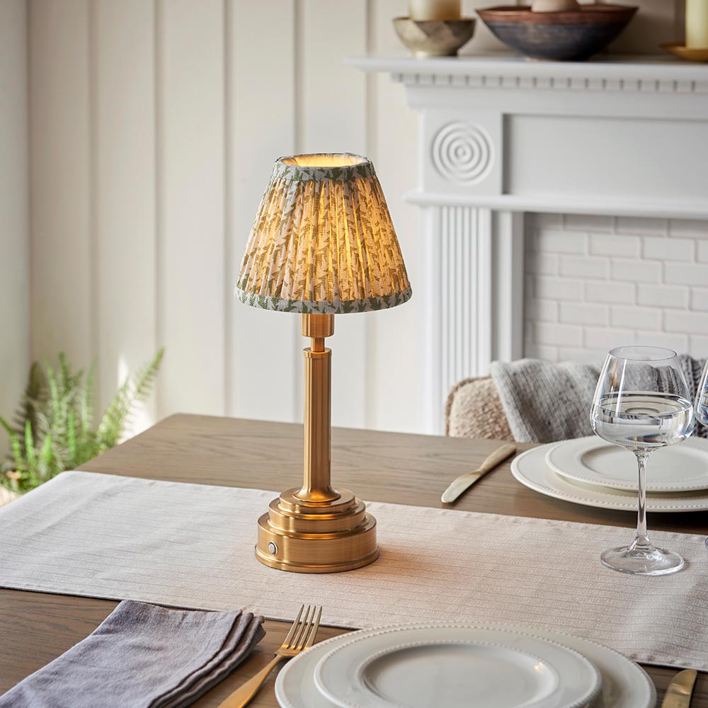 Endon Danebourne LED Leaf table lamp with brushed brass stepped base and green leaf-print pleated shade, lit on a dining table beside wine glasses.