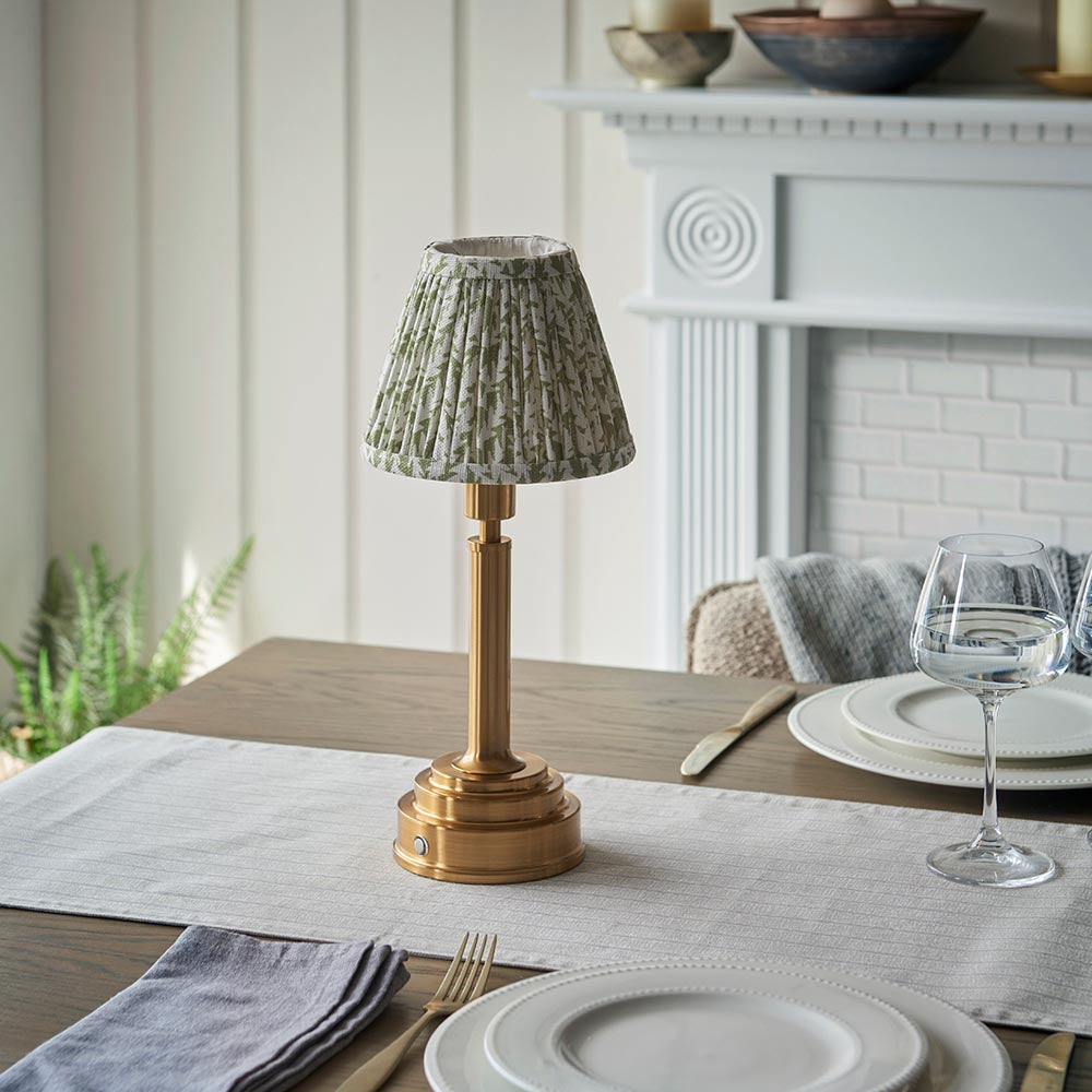 Endon Danebourne LED table lamp with brushed brass stepped base and green leaf-print pleated fabric shade, styled on a dining table setting.