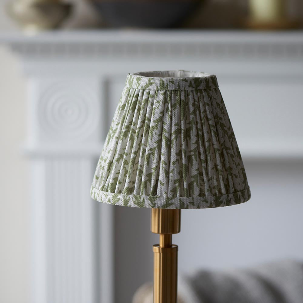 Endon Danebourne LED Leaf table lamp with a pleated green and white faux linen shade on a brushed aged brass stepped base, shown by a pale fireplace.