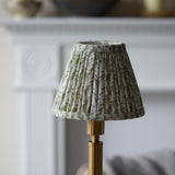 Endon Danebourne LED Leaf table lamp with a pleated green and white faux linen shade on a brushed aged brass stepped base, shown by a pale fireplace.