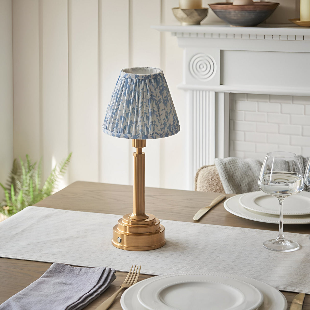 Danebourne LED Leaf Table Lamp in brass with a pleated blue and white leaf-print fabric shade, shown on a dining table near a white fireplace mantel.