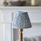 Danebourne LED Leaf Table Lamp with pleated white and blue leaf-print fabric shade on a brass stepped base, shown against a soft-focus mantelpiece setting.
