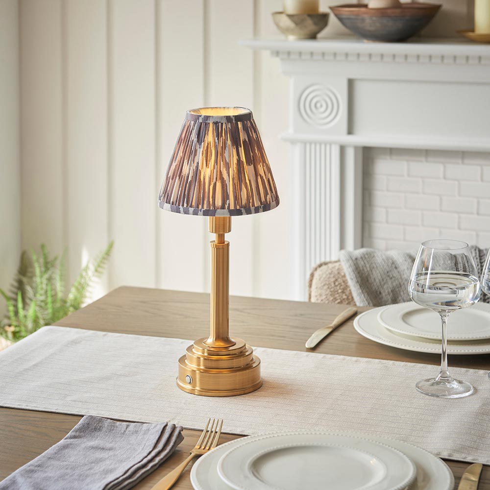 Endon Danebourne LED Ikat table lamp with brushed brass stepped base and grey pleated faux silk shade, lit on a dining table beside wine glasses.