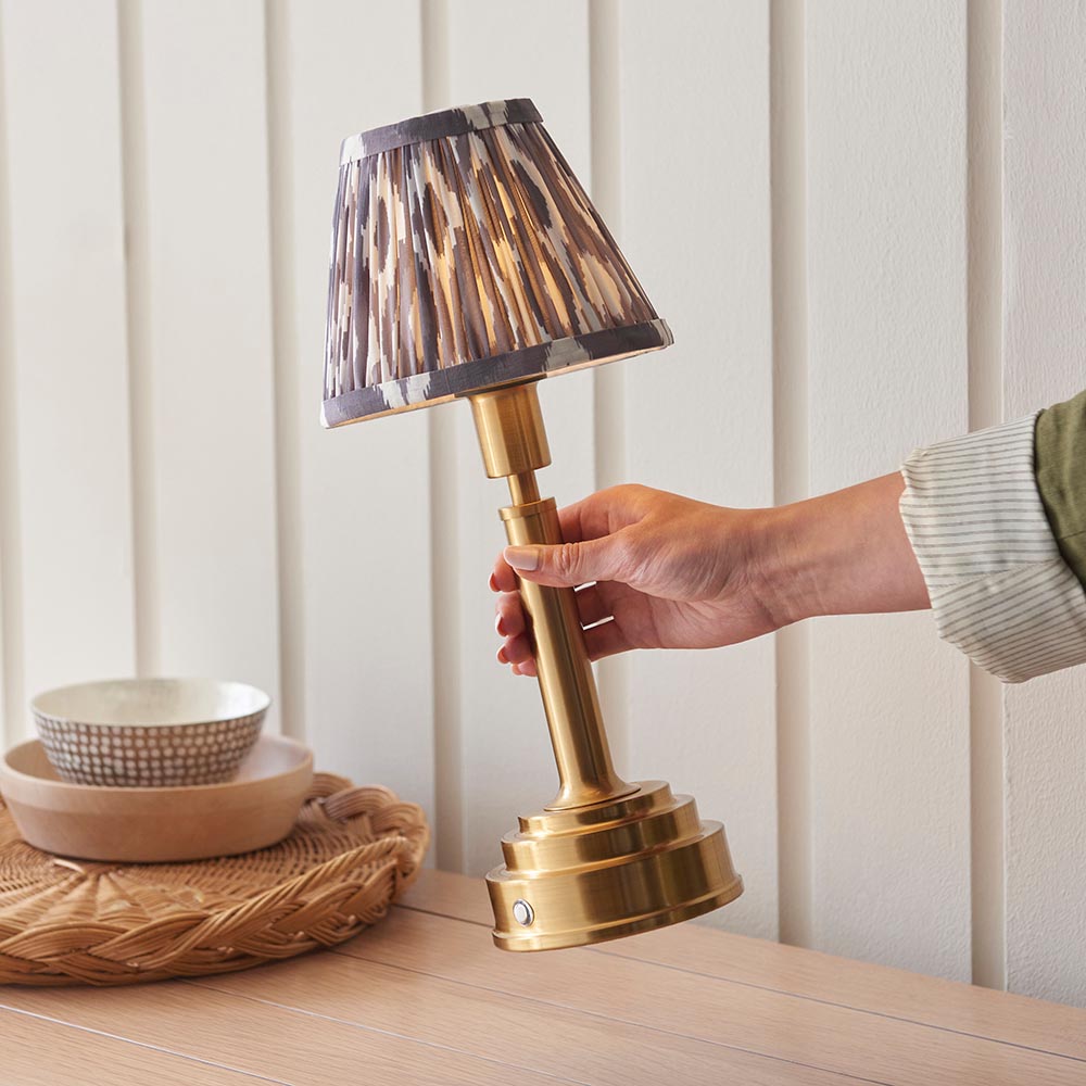 Hand adjusting the Endon Danebourne LED Ikat table lamp with brushed brass stepped base and grey and white Ikat clip-on shade on a wooden sideboard.