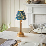 Danebourne LED Ikat Table Lamp with brass stepped base and blue patterned fabric shade, shown lit on a dining table near a white fireplace.