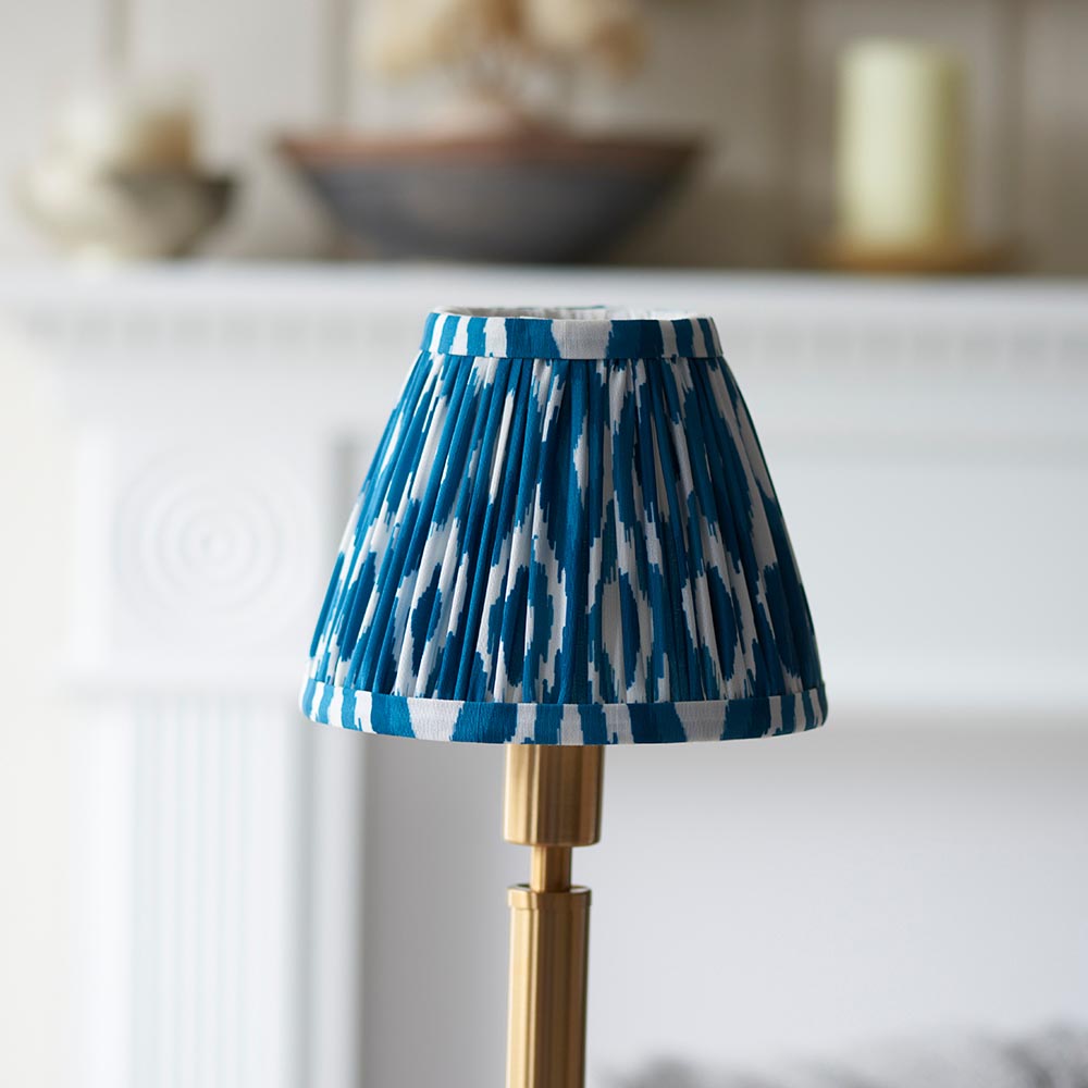 Danebourne LED Ikat Table Lamp with brass stepped base and pleated blue and white patterned shade, shown on a mantelpiece in a living room setting.