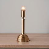 Danebourne LED table lamp with stepped brass column base, exposed globe bulb, and touch control, shown lit on wood surface against a plain grey background.