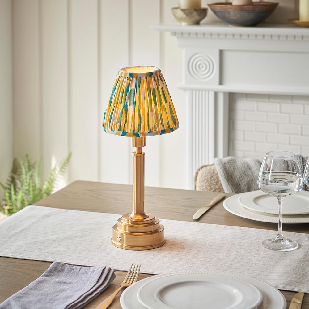 Endon Danebourne LED Ikat table lamp with brushed brass stepped base and yellow, white and teal pleated shade, lit on a dining table setting.