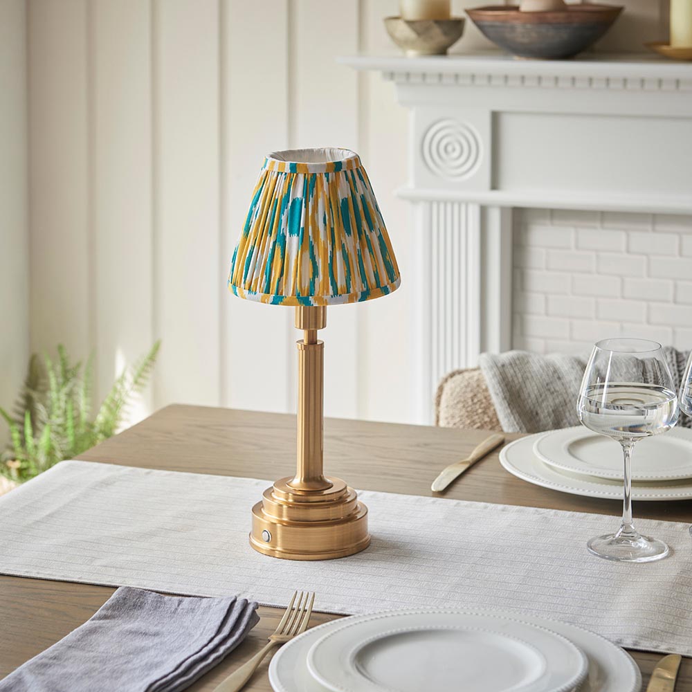 Endon Danebourne LED Ikat table lamp with brushed brass stepped base and yellow, teal and white pleated fabric shade on a dining table.