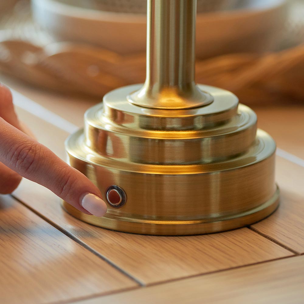 Finger pressing the touch dimmer on the brushed brass stepped base of the Endon Danebourne LED Ikat Table Lamp on a wooden surface.