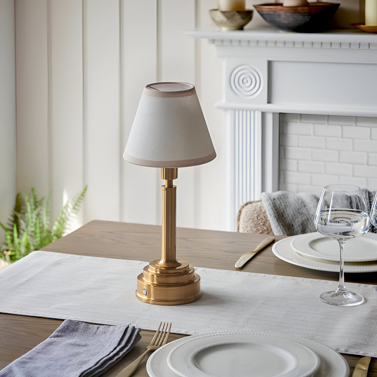 Danebourne LED rechargeable table lamp in brass with cream fabric shade, styled on a dining table beside white plates and a glass, near a white fireplace.